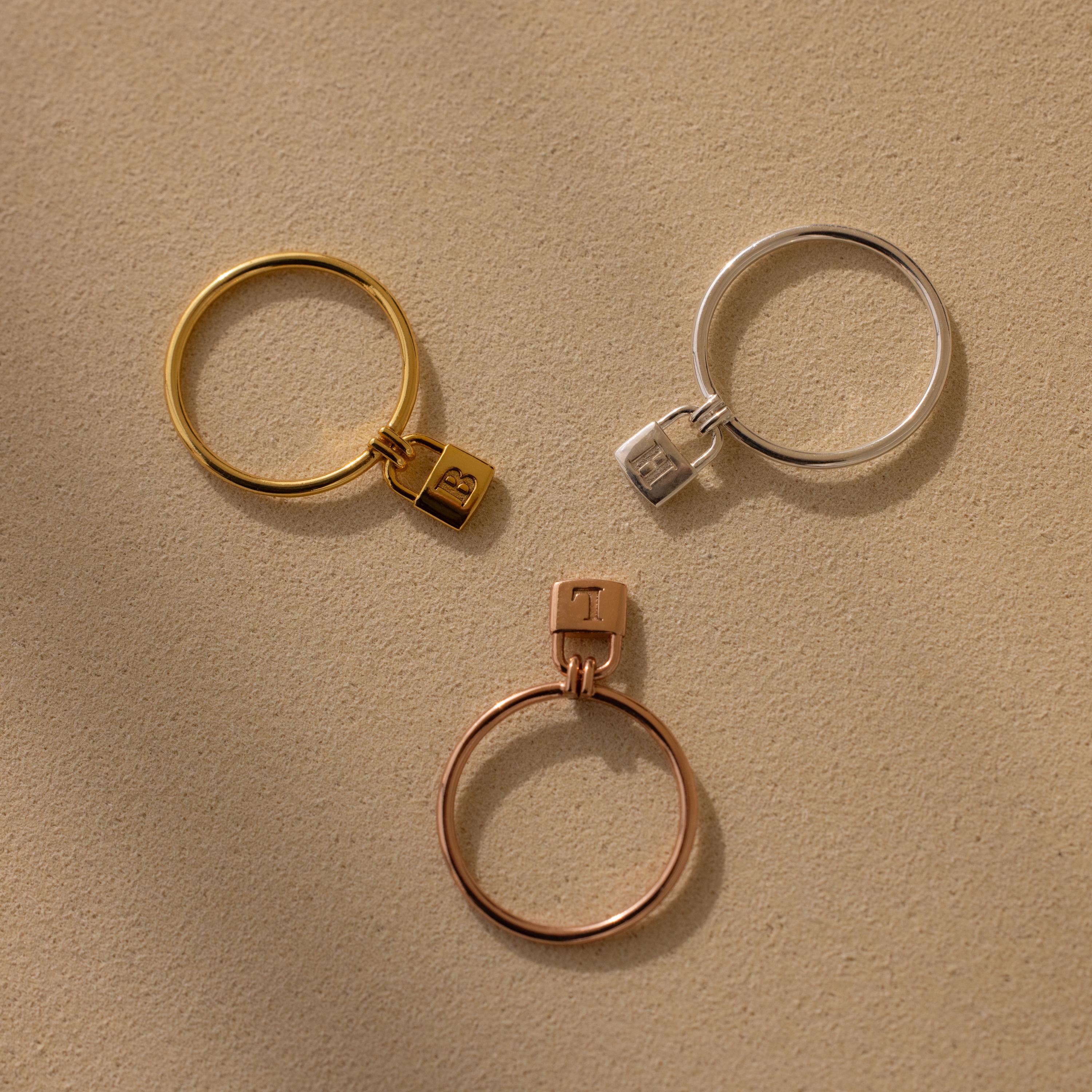Three Initial Padlock Rings in different finishes: gold, silver, and rose gold, each with a padlock charm on a beige background.